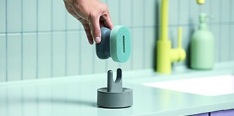 Mano colocando un estropajo de mano Brite de Scotch-Brite™ en un soporte para estropajos sobre la encimera de la cocina, junto al fregadero.
